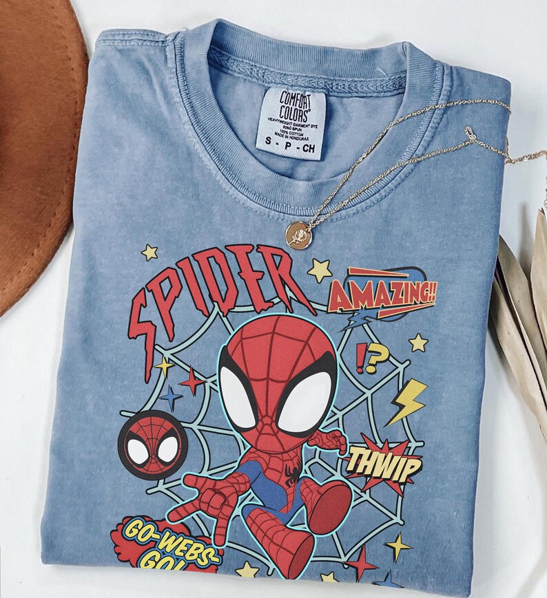 May include: A blue short-sleeved t-shirt featuring a graphic of a cartoon spider character in a red and blue suit. The design includes a spider web background, yellow stars, and text that reads SPIDER, AMAZING, THWIP, and GO-WEBS-GO.