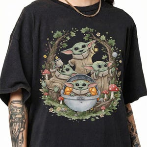 May include: A black short-sleeve t-shirt featuring a graphic illustration of four green creatures with large ears in a forest setting. One creature sits in a floating pod, while others hold a lantern and a mug among trees, leaves, and red mushrooms.
