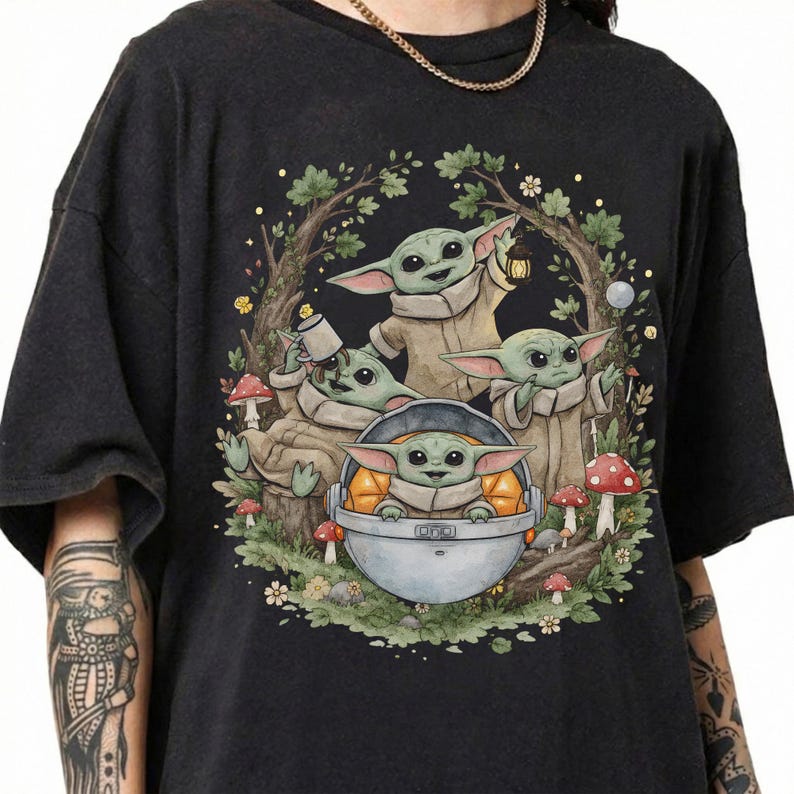 May include: A black short-sleeve t-shirt featuring a graphic illustration of four green creatures with large ears in a forest setting. One creature sits in a floating pod, while others hold a lantern and a mug among trees, leaves, and red mushrooms.