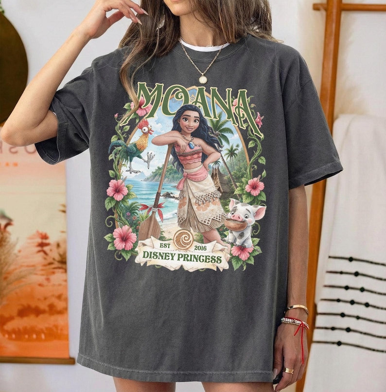 May include: A charcoal gray short-sleeve t-shirt featuring a graphic print of the character Moana, a rooster, and a pig. The design includes tropical flowers, a wooden oar, and the text MOANA and EST 2016 DISNEY PRINCESS in a decorative frame.