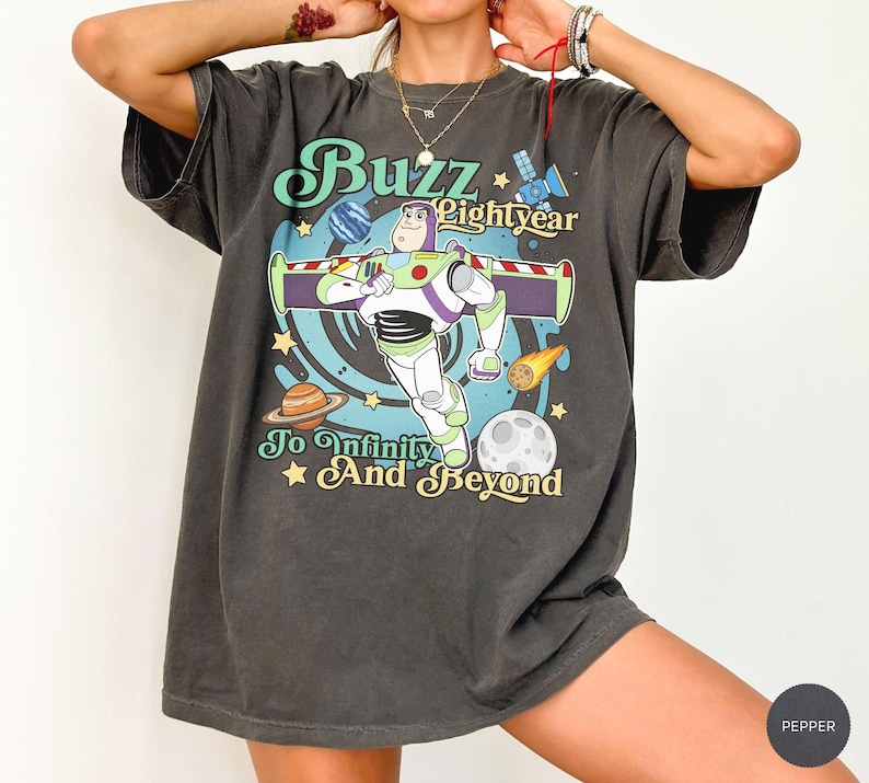 May include: A charcoal gray short-sleeve t-shirt featuring a graphic of Buzz Lightyear in space with planets, stars, and a satellite. The text reads Buzz Lightyear To Infinity And Beyond in a retro font style with light blue and yellow accents.