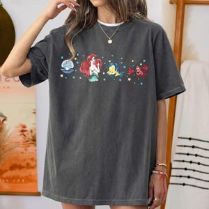 K&ouml;nnte beinhalten: Dunkelgraues T-Shirt mit einem farbenfrohen Aufdruck von Zeichentrickfiguren. Das Design umfasst eine Meerjungfrau, einen Fisch, eine Krabbe und eine Muschel. Das Shirt hat eine lockere Passform und kurze &Auml;rmel.