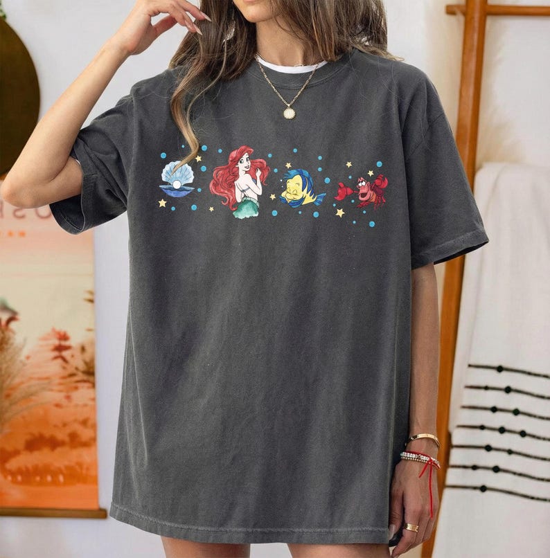 K&ouml;nnte beinhalten: Dunkelgraues T-Shirt mit einem farbenfrohen Aufdruck von Zeichentrickfiguren. Das Design umfasst eine Meerjungfrau, einen Fisch, eine Krabbe und eine Muschel. Das Shirt hat eine lockere Passform und kurze &Auml;rmel.