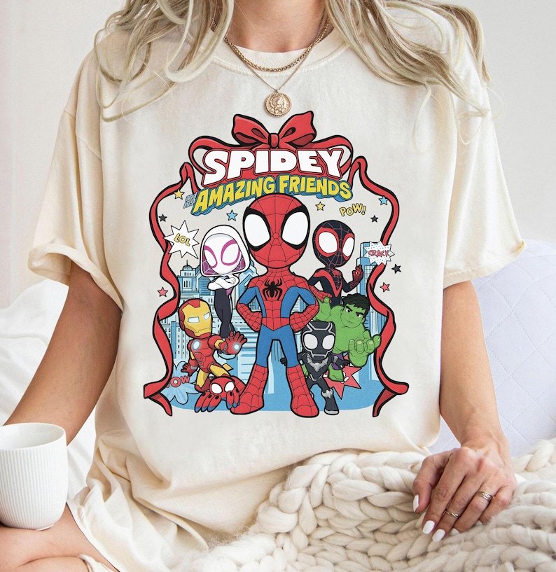 May include: A cream-coloured t-shirt featuring a graphic print of Spidey and His Amazing Friends. The design includes cartoon versions of Spider-Man, Ghost-Spider, Miles Morales, Iron Man, Black Panther, and Hulk framed by a red ribbon.