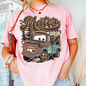May include: Pink t-shirt featuring a cartoon image of Mater from the movie Cars. The graphic includes the text "Mater 2026" in a brown font. The design is set against a backdrop of a forest.
