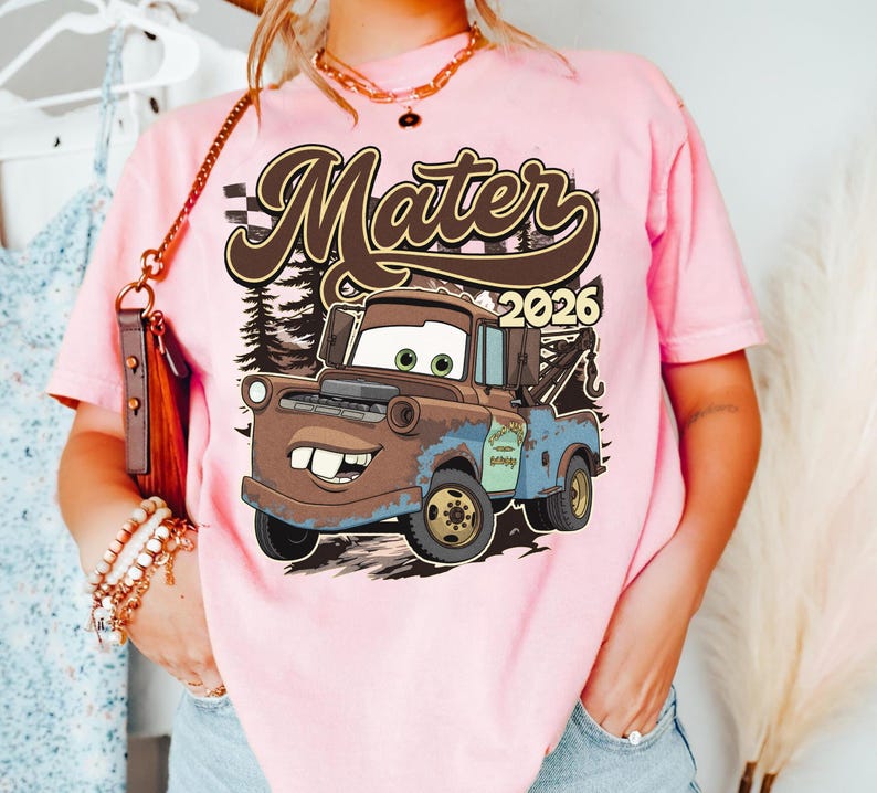 May include: Pink t-shirt featuring a cartoon image of Mater from the movie Cars. The graphic includes the text "Mater 2026" in a brown font. The design is set against a backdrop of a forest.