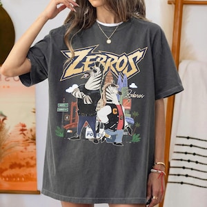 May include: A charcoal grey short-sleeve t-shirt featuring a graphic print of two anthropomorphic zebras in a city setting. The text "ZEBROS" appears in large yellow letters at the top, with "Zekaros" written in script below the zebras.