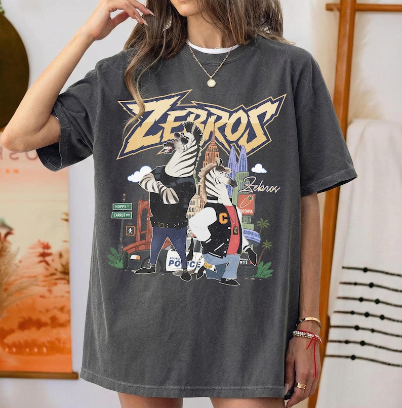 May include: A charcoal grey short-sleeve t-shirt featuring a graphic print of two anthropomorphic zebras in a city setting. The text "ZEBROS" appears in large yellow letters at the top, with "Zekaros" written in script below the zebras.