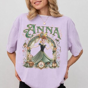May include: A light purple short-sleeve t-shirt featuring a graphic print of a character in a green dress standing under an archway. The text ANNA is printed in large green letters above the character, surrounded by floral and bird illustrations.