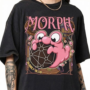 Puede incluir: Una camiseta negra de manga corta con un estampado gráfico de una criatura rosa de dibujos animados que sostiene una esfera oscura con un patrón de líneas geométricas blancas. El texto "MORPH" aparece en rosa arriba y "BUBBLY BLOB" en amarillo abajo.