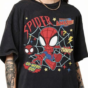 May include: A black t-shirt featuring a graphic of a cartoon Spider-Man character in a web. The design includes the text "SPIDER", "AMAZING!!", "THWIP", and "GO-WEBS GO!" along with yellow stars, a lightning bolt, and a question mark exclamation point.