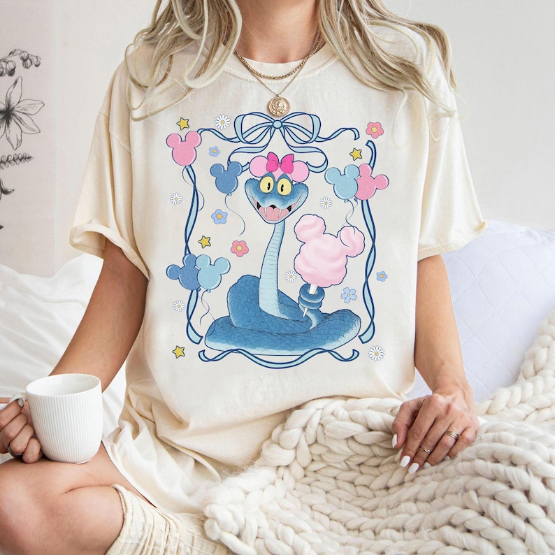 May include: Cream-colored t-shirt featuring a cartoon blue snake with a pink bow, holding cotton candy. The design includes pink and blue Mickey Mouse-shaped balloons and flowers, framed by a blue border.