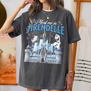 May include: Dark gray t-shirt featuring a graphic of a castle, reindeer, and snowman with the text "Meet me in Arendelle" in blue and white. The shirt has a relaxed fit and short sleeves.