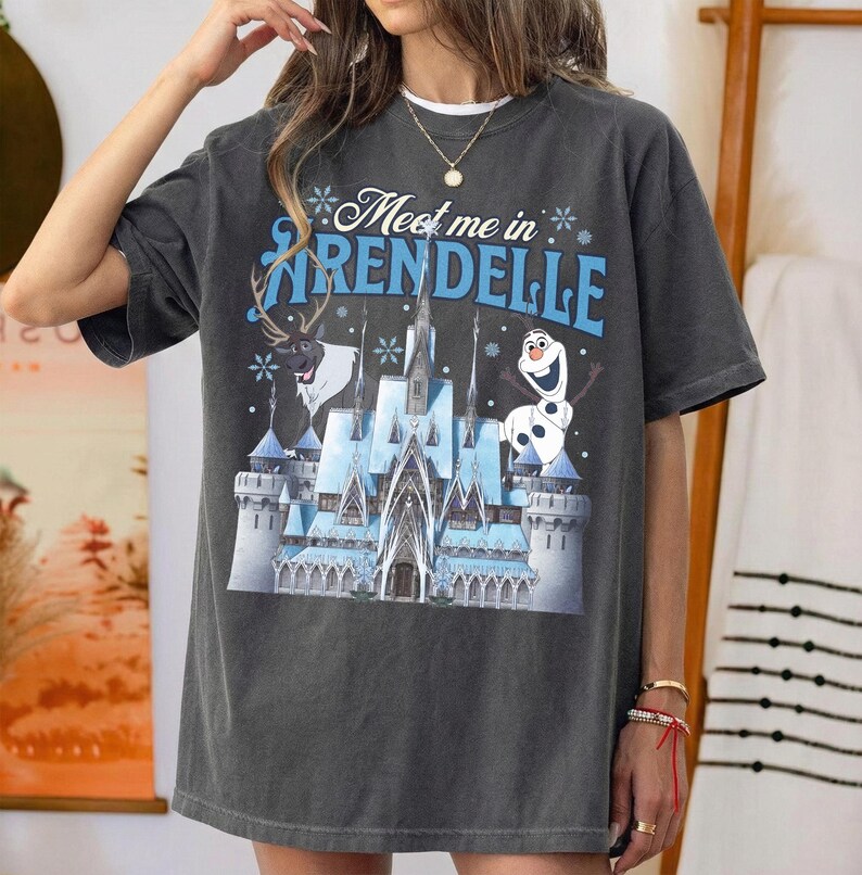 May include: Dark gray t-shirt featuring a graphic of a castle, reindeer, and snowman with the text "Meet me in Arendelle" in blue and white. The shirt has a relaxed fit and short sleeves.