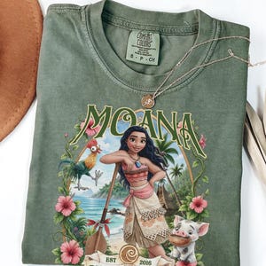 May include: An olive green crew neck t-shirt featuring a graphic print of the character Moana, a rooster, and a pig. The design includes the text MOANA, EST 2016, and tropical floral accents. The shirt has a Comfort Colors brand label inside.