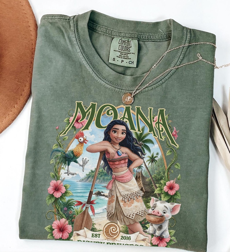 May include: An olive green crew neck t-shirt featuring a graphic print of the character Moana, a rooster, and a pig. The design includes the text MOANA, EST 2016, and tropical floral accents. The shirt has a Comfort Colors brand label inside.