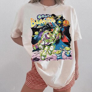 Puede incluir: Una camiseta de color crema con un estampado gr&aacute;fico de Buzz Lightyear y alien&iacute;genas verdes de Astro Blasters. El dise&ntilde;o incluye el texto Astro Blasters en letras amarillas y azules, sobre un fondo espacial oscuro con planetas.