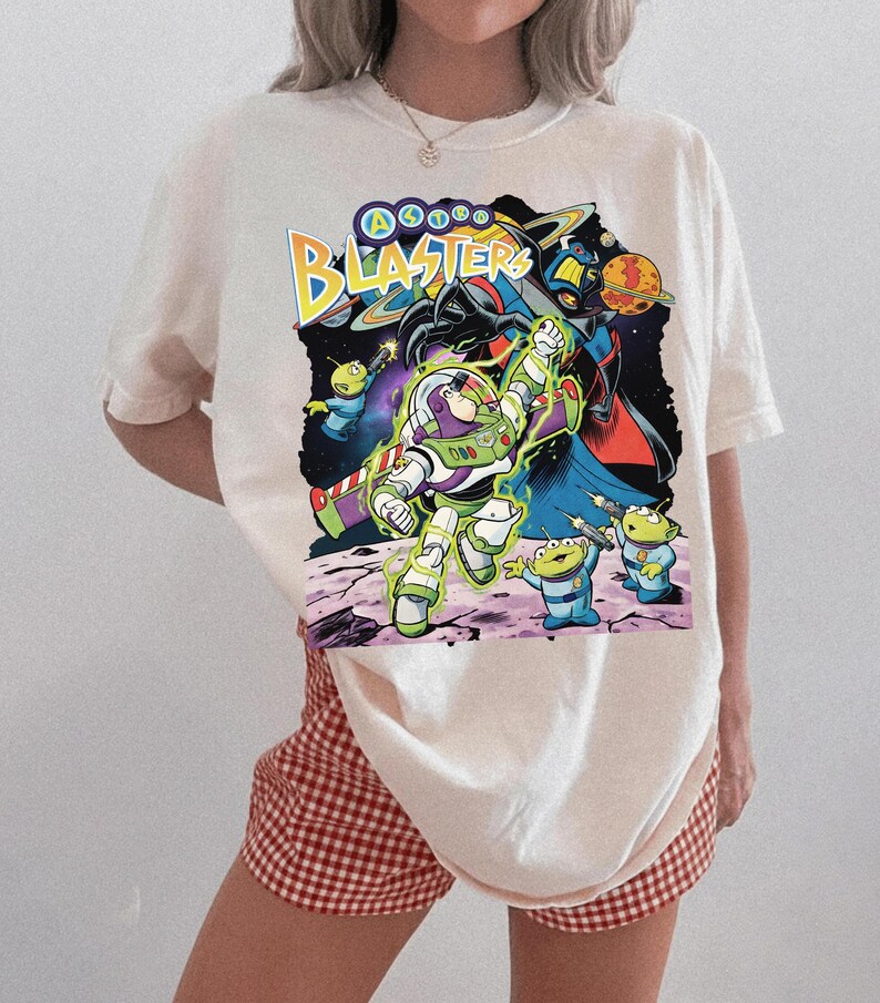 Puede incluir: Una camiseta de color crema con un estampado gr&aacute;fico de Buzz Lightyear y alien&iacute;genas verdes de Astro Blasters. El dise&ntilde;o incluye el texto Astro Blasters en letras amarillas y azules, sobre un fondo espacial oscuro con planetas.