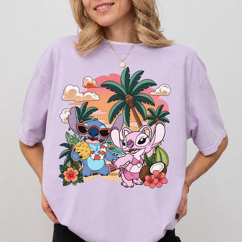 May include: A light purple t-shirt featuring a graphic print of two cartoon characters on a beach. One blue character wears red sunglasses and holds a pineapple drink, while a pink character stands beside it. Palm trees and hibiscus flowers surround them.