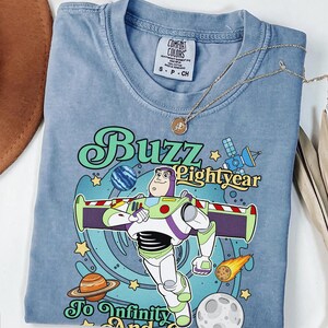 May include: A blue short-sleeve t-shirt featuring a graphic of Buzz Lightyear with the text "Buzz Lightyear To Infinity And Beyond". The design includes planets, a satellite, and a comet against a swirling blue background. A tag reads Comfort Colors.