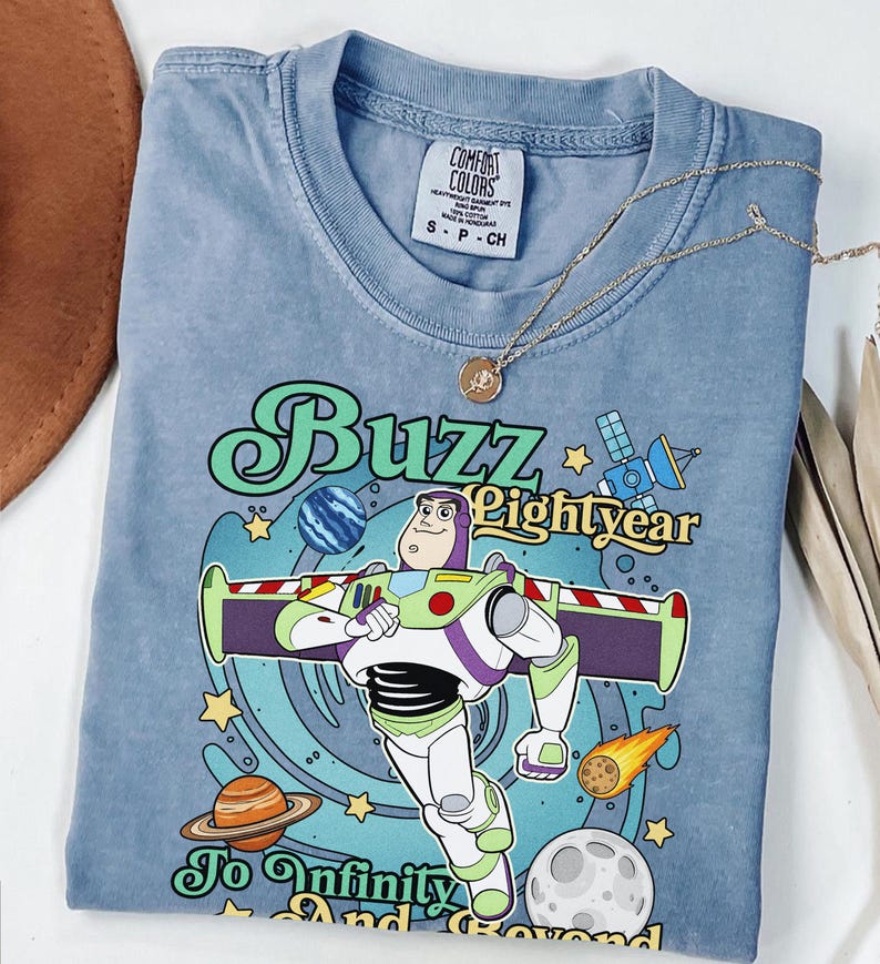 May include: A blue short-sleeve t-shirt featuring a graphic of Buzz Lightyear with the text "Buzz Lightyear To Infinity And Beyond". The design includes planets, a satellite, and a comet against a swirling blue background. A tag reads Comfort Colors.