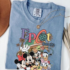 May include: A light blue Comfort Colors t-shirt featuring the word "Epcot" in colorful lettering with a globe graphic. The shirt includes the text "Since 1982" and a graphic of Disney characters. A gold necklace with a pendant rests on the shirt.