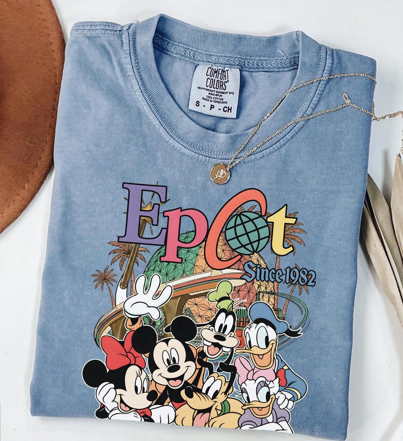 May include: A light blue Comfort Colors t-shirt featuring the word "Epcot" in colorful lettering with a globe graphic. The shirt includes the text "Since 1982" and a graphic of Disney characters. A gold necklace with a pendant rests on the shirt.