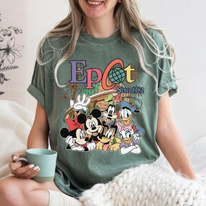 May include: Green t-shirt featuring the word "Epcot" in colorful letters with a globe graphic and the text "Since 1982." The shirt also displays cartoon characters Mickey Mouse, Minnie Mouse, Goofy, Donald Duck, and Daisy Duck.