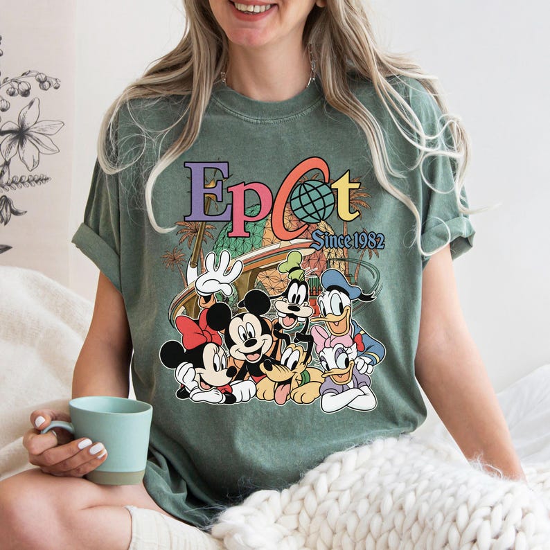 May include: Green t-shirt featuring the word "Epcot" in colorful letters with a globe graphic and the text "Since 1982." The shirt also displays cartoon characters Mickey Mouse, Minnie Mouse, Goofy, Donald Duck, and Daisy Duck.