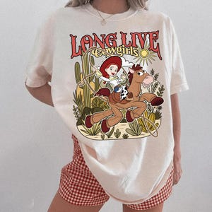 Puede incluir: Una camiseta color hueso con un estampado gr&aacute;fico de una vaquera de dibujos animados montando un caballo marr&oacute;n con un lazo. El texto "LONG LIVE cowgirls" est&aacute; impreso en rojo sobre la ilustraci&oacute;n, que incluye cactus, un sol y vegetaci&oacute;n.