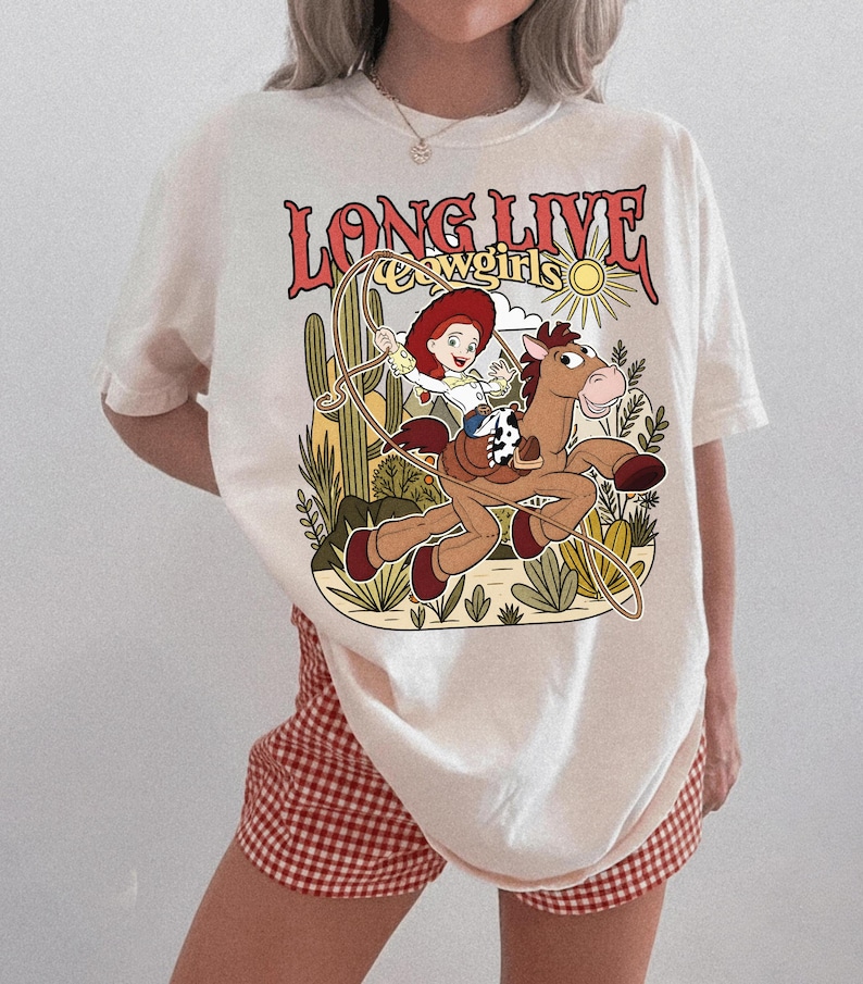 Puede incluir: Una camiseta color hueso con un estampado gr&aacute;fico de una vaquera de dibujos animados montando un caballo marr&oacute;n con un lazo. El texto "LONG LIVE cowgirls" est&aacute; impreso en rojo sobre la ilustraci&oacute;n, que incluye cactus, un sol y vegetaci&oacute;n.
