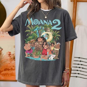 May include: A charcoal gray short-sleeve t-shirt featuring a graphic print of characters from the movie Moana 2. The design includes a waterfall, palm trees, and the text Moana 2 in light blue. The shirt has a crew neckline and a relaxed fit.