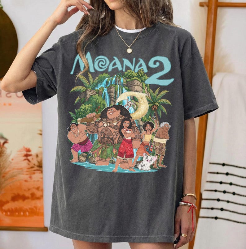May include: A charcoal gray short-sleeve t-shirt featuring a graphic print of characters from the movie Moana 2. The design includes a waterfall, palm trees, and the text Moana 2 in light blue. The shirt has a crew neckline and a relaxed fit.