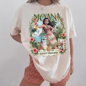 May include: An off-white short-sleeve t-shirt featuring a graphic print of Moana, a rooster, and a piglet. The design includes tropical flowers, a wooden oar, and the text "MOANA" at the top and "EST 2016 DISNEY PRINCESS" at the bottom.