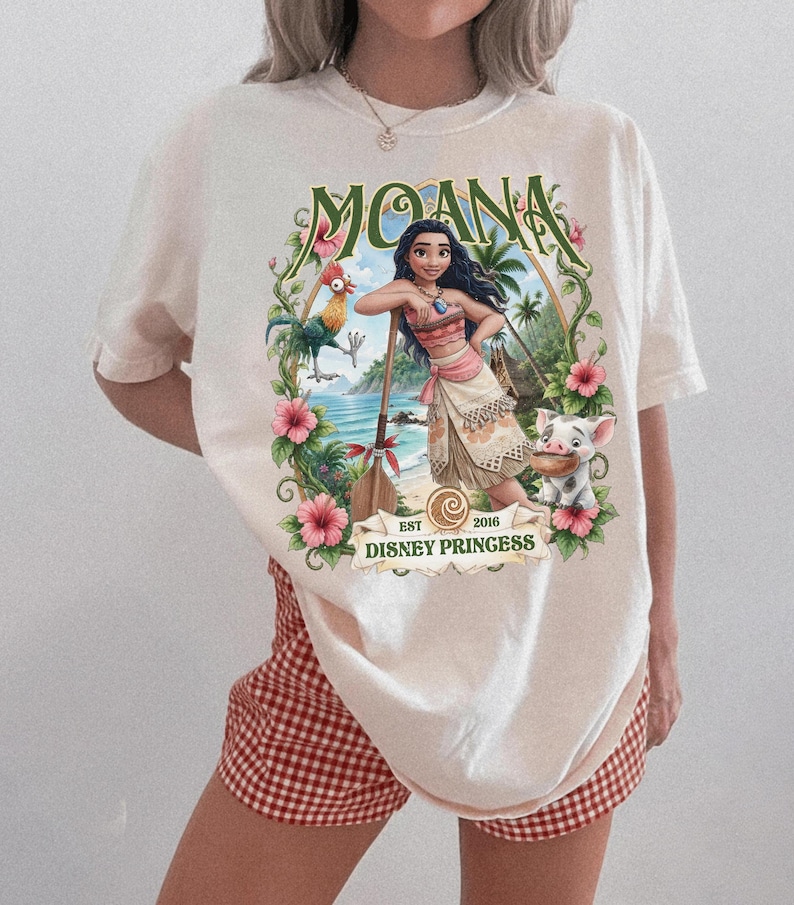May include: An off-white short-sleeve t-shirt featuring a graphic print of Moana, a rooster, and a piglet. The design includes tropical flowers, a wooden oar, and the text "MOANA" at the top and "EST 2016 DISNEY PRINCESS" at the bottom.