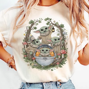 May include: A cream-colored t-shirt featuring a graphic illustration of four green, large-eared creatures in a forest setting. One creature sits in a floating silver pod, while others interact with a lantern, a mug, and red-spotted mushrooms.