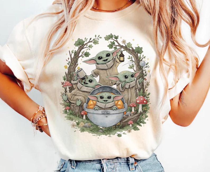 May include: A cream-colored t-shirt featuring a graphic illustration of four green, large-eared creatures in a forest setting. One creature sits in a floating silver pod, while others interact with a lantern, a mug, and red-spotted mushrooms.