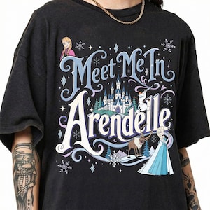 K&ouml;nnte beinhalten: Ein schwarzes T-Shirt mit einem Grafikdruck, der den Text "Meet Me In Arendelle" in stilisierter blauer und wei&szlig;er Schrift zeigt. Das Design enth&auml;lt Illustrationen von Anna, Elsa, Olaf und Sven sowie ein Schloss und Schneeflocken.