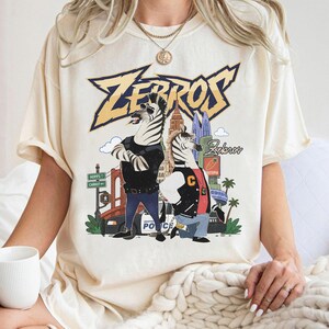 May include: A cream-coloured short-sleeve t-shirt featuring a graphic print of two anthropomorphic zebras in a city setting. The text "ZEBROS" appears in large yellow letters at the top, with a police car, city buildings, and street signs visible.