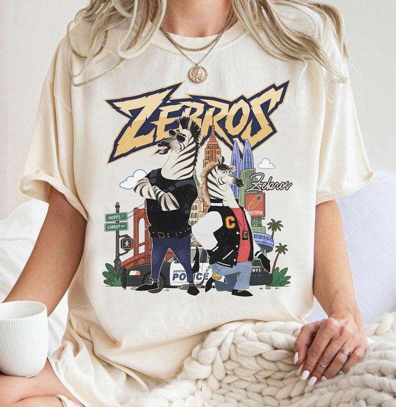 May include: A cream-coloured short-sleeve t-shirt featuring a graphic print of two anthropomorphic zebras in a city setting. The text "ZEBROS" appears in large yellow letters at the top, with a police car, city buildings, and street signs visible.