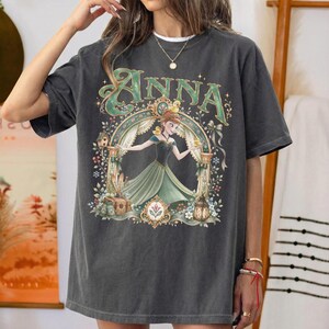 May include: A charcoal gray short-sleeve t-shirt featuring a graphic print of a character in a green dress under an archway. The text ANNA is printed in large green letters above the character. The design includes floral accents and small birds.