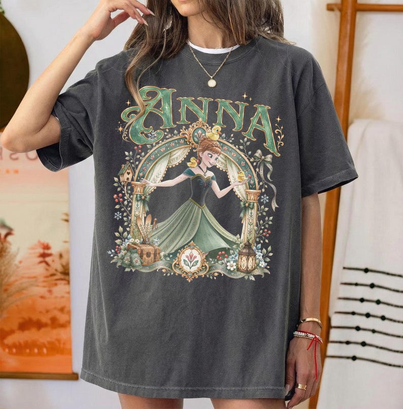 May include: A charcoal gray short-sleeve t-shirt featuring a graphic print of a character in a green dress under an archway. The text ANNA is printed in large green letters above the character. The design includes floral accents and small birds.