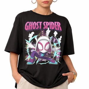 May include: Black t-shirt featuring a Ghost Spider graphic. The design includes a cartoon illustration of Ghost Spider in a dynamic pose, set against a cityscape background. The text "Ghost Spider" is displayed in pink and purple.