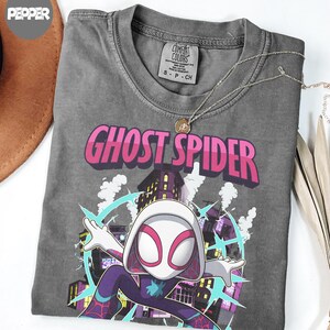 May include: A gray t-shirt featuring the words "GHOST SPIDER" in pink and white, with a graphic of a superhero in a cityscape. The shirt has a crew neck and short sleeves. A silver necklace with a pendant is on the shirt.