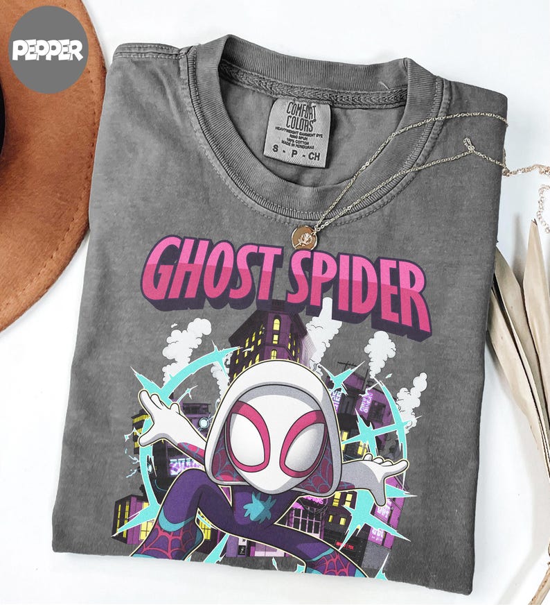 May include: A gray t-shirt featuring the words "GHOST SPIDER" in pink and white, with a graphic of a superhero in a cityscape. The shirt has a crew neck and short sleeves. A silver necklace with a pendant is on the shirt.