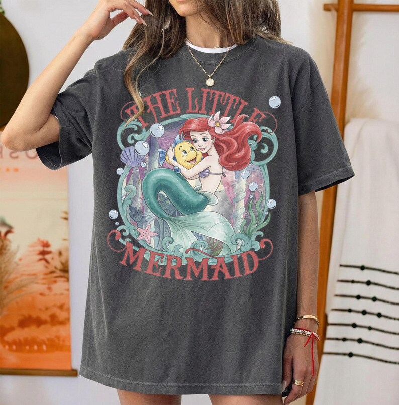 May include: A charcoal gray short-sleeve t-shirt featuring a graphic print of Ariel and Flounder from The Little Mermaid. The design includes red text reading THE LITTLE MERMAID, surrounded by teal seaweed, bubbles, a seashell, and a starfish.