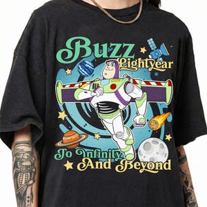 May include: A black t-shirt featuring a graphic of Buzz Lightyear in a space setting with planets, stars, and a satellite. The text reads Buzz Lightyear To Infinity And Beyond in a stylized font, surrounded by a blue swirling cosmic background.