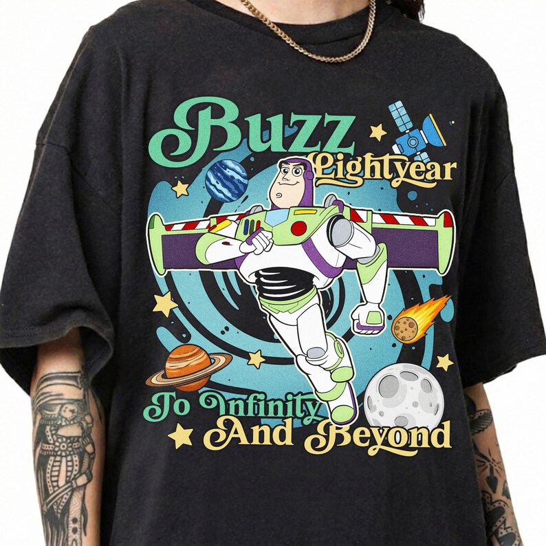 May include: A black t-shirt featuring a graphic of Buzz Lightyear in a space setting with planets, stars, and a satellite. The text reads Buzz Lightyear To Infinity And Beyond in a stylized font, surrounded by a blue swirling cosmic background.