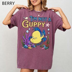Puede incluir: Camiseta color bayas con el texto "I'm Not a Guppy" y una imagen de dibujos animados de Flounder de La Sirenita. El gr&aacute;fico incluye coral, burbujas y una estrella de mar. La camiseta es de corte holgado.