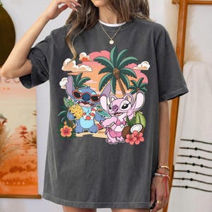 May include: A charcoal gray crew neck t-shirt featuring a graphic print of two cartoon characters, one blue and one pink, standing on a beach with palm trees, hibiscus flowers, and a pineapple drink against a sunset background with clouds.